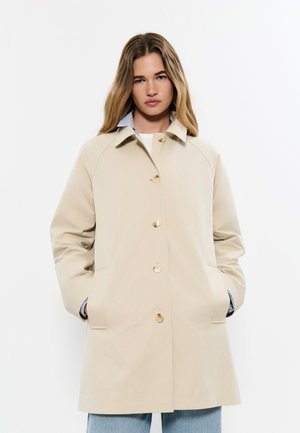 Mujer con abrigo beige abotonado y manos en los bolsillos, con vaqueros azul claro, sobre un fondo blanco liso.