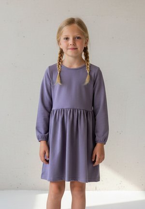 Młoda dziewczyna z blond warkoczem, ubrana w lawendową sukienkę z długim rękawem, stojąca na tle jednolitej jasnej ściany.