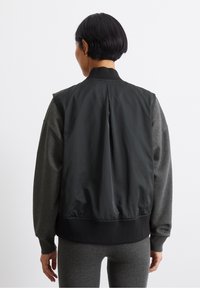 Zwarte nylon bomberjack met grijze gebreide mouwen en geribde kraag; heeft een geplooide achterkant en een elastische tailleband voor een ontspannen pasvorm.