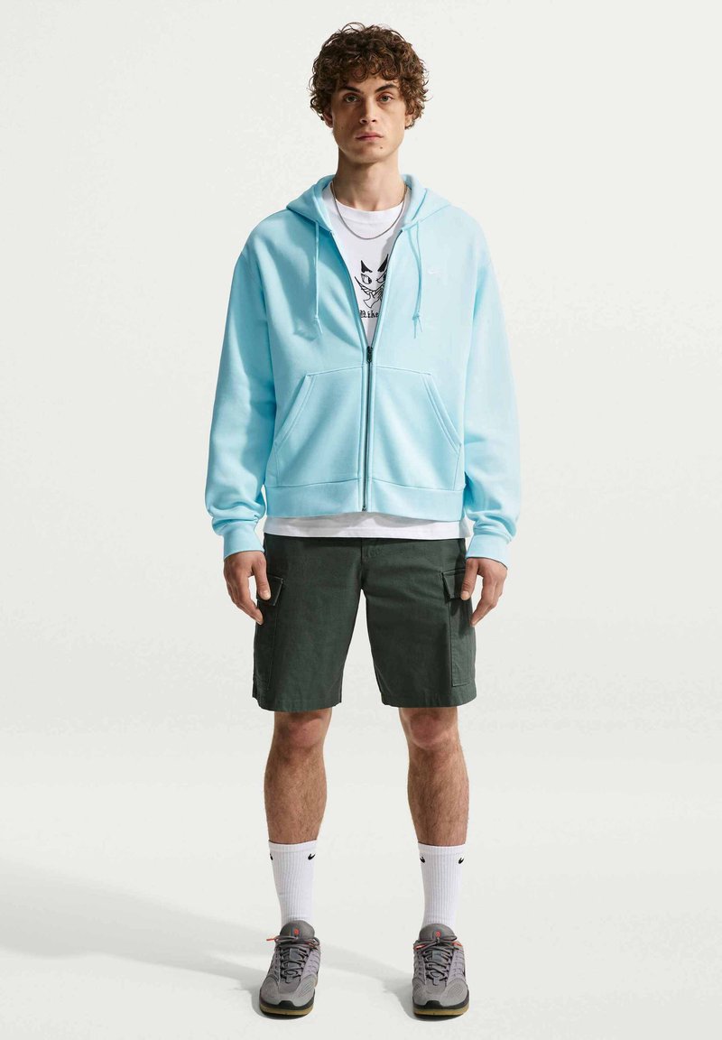 Νέος άνδρας που στέκεται φορώντας ανοιχτό μπλε φούτερ με φερμουάρ, λευκό μπλουζάκι με  στάμπα, σκούρα cargo σορτς, λευκές κάλτσες και γκρι αθλητικά σε απλό φόντο.