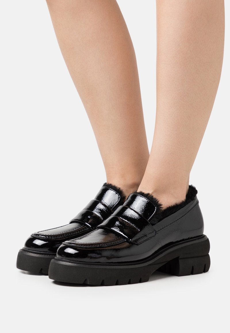 Mocassins en cuir verni noir avec semelles épaisses et doublure en fourrure noire portés sur des jambes nues sur fond blanc.