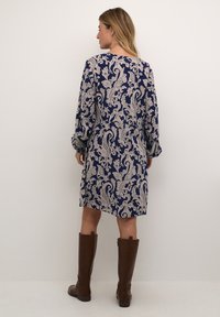 Robe à manches longues en bleu marine avec un motif paisley beige, fabriquée en tissu lisse. Associée à des bottes en cuir marron jusqu'au genou.