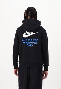 Person med flätat hår som bär en svart Nike-hoodie med texten "BARA AUTORISERADE PERSONER" i blått på ryggen mot en enfärgad vit bakgrund.