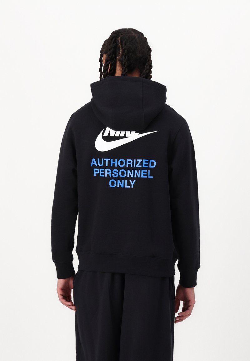 Person med flätat hår som bär en svart Nike-hoodie med texten "BARA AUTORISERADE PERSONER" i blått på ryggen mot en enfärgad vit bakgrund.