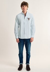 Camisa de botón a rayas azul claro con mangas largas, que presenta un logo bordado, combinada con jeans oscuros y zapatos de ante marrones.