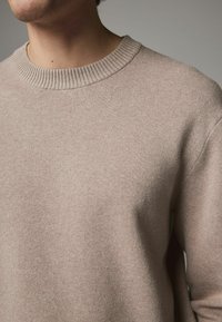 Beżowy sweter z okrągłym dekoltem o teksturowanym wzorze. Kołnierz posiada ryżowy detal. Materiał wydaje się miękki i lekki.