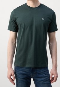 Man in een donkergroene T-shirt met korte mouwen en ronde hals, voorzien van een klein wit "MOP" logo op de linkerkant van de borst, gecombineerd met blauwe spijkerbroek.