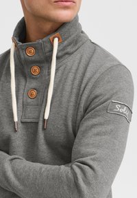 Pilkas megztinis su aukštu apykakliu, mediniais mygtukais, kreminės spalvos raišteliais ir audinio lopu ant rankovės su užrašu „Solus“.