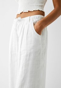Białe lniane spodnie o luźnym kroju, z elastycznym pasem i bocznymi kieszeniami. Materiał ma teksturowany, lekko pomarszczony wygląd.