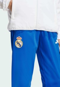 Blaue Sporthosen mit strukturierter Oberfläche, ausgestattet mit weißen Adidas-Streifen und einem Vereinslogo mit einer Krone auf dem Oberschenkel.