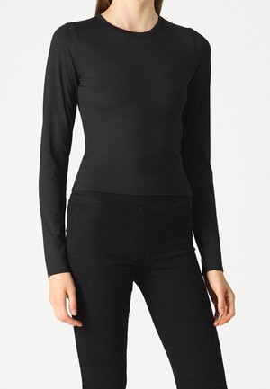 Mujer vestida con una camiseta negra ajustada de manga larga y pantalones negros, de pie frente a un fondo blanco liso.