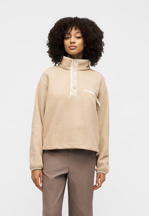 Mujer con cabello rizado que lleva un pulóver de forro polar beige con botones de presión y un bolsillo con cremallera en el pecho, combinado con pantalones marrones.