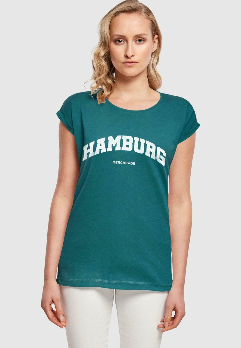 Tyrkysové tričko s krátkým rukávem s nápisem "HAMBURG" v bílém zakřiveném písmu, vyrobené z měkké bavlny. Design zahrnuje zaoblený výstřih a uvolněný střih.