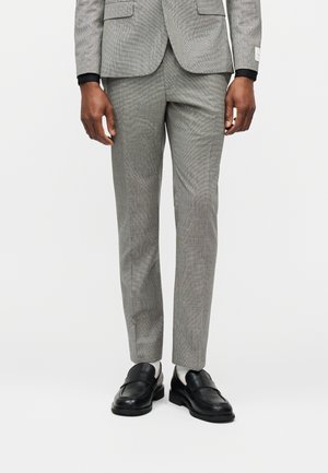 Man wearing slim-fit grey suit trousers, black loafers, and white socks, standing against a plain white background.