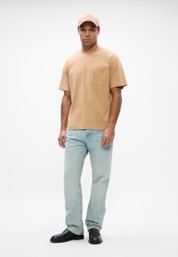 Μπεζ κοντομάνικο μπλουζάκι με τσέπη στην επάνω πλευρά, συνδυασμένο με ανοιχτό μπλε ίσια τζιν παντελόνια και μαύρα παπούτσια. Καπέλο με το λογότυπο "BOSS" εμπρός.