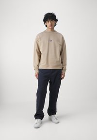 Felpa beige con spalle scese, polsini a costine e un piccolo logo blu; abbinata a pantaloni blu e sneaker bianche. Sfondo semplice.