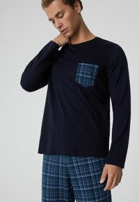 Camisa de manga larga azul marino con un bolsillo a cuadros de color teal, combinada con pantalones cortos de cuadros en tonos de azul y verde, hecha de una tela suave.