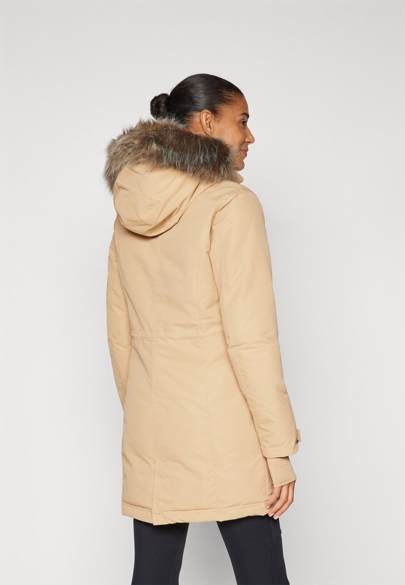 Parka beige avec une capuche bordée de fourrure, présentant une coupe ajustée, des manches longues et une longueur mi-cuisse pour la chaleur et la couverture.
