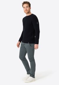 Marinefarbener Strickpullover mit strukturiertem Muster, kombiniert mit schmal geschnittenen grünen Hosen und weißen Sneakers. Detaillierte Nähte und Logo auf dem Ärmel.