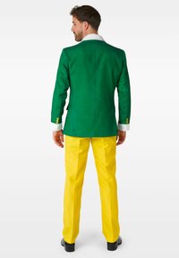 Blazer verde con colletto bianco, abbinato a pantaloni gialli. L'outfit presenta bottoni dorati sui polsini e una vestibilità sartoriale.