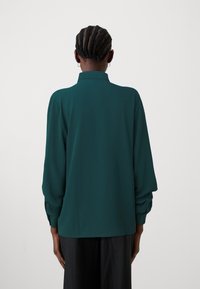 Bluză teal cu guler înalt, mâneci lungi cu mansete plisate și un material neted, texturat. Accente subtile cu nasturi la mansete.