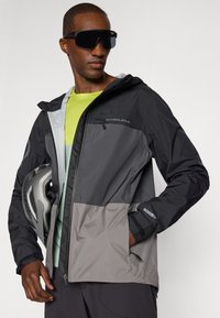 Veste imperméable noire et grise avec fermeture éclair à l'avant, capuche ajustable et grande poche avant. Comprend des accents réfléchissants et des poignets élastiques.