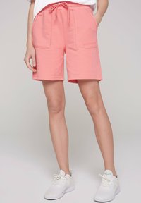 Shorts di cotone rosa con vita regolabile, due tasche frontali e dettaglio del logo. Indossati con scarpe da ginnastica bianche. Texture liscia.