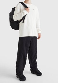 Bílá dlouhá košile s dlouhým rukávem, černý batoh, tmavé džíny a černé boty. Obsahuje žebrovanou strukturu a detail loga na košili.