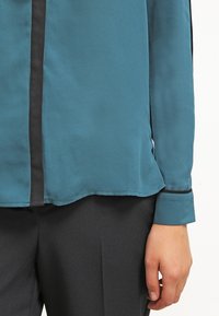 Turkos blus med svart kant, med en jämn textur, långa ärmar och en något böjd fåll. Bärs med mörka byxor.