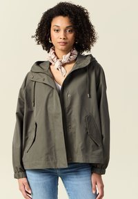 Giacca impermeabile verde oliva con cappuccio, zip frontale, due tasche laterali e polsini elasticizzati. Indossata sopra un top leggero e accessoriate con una sciarpa.