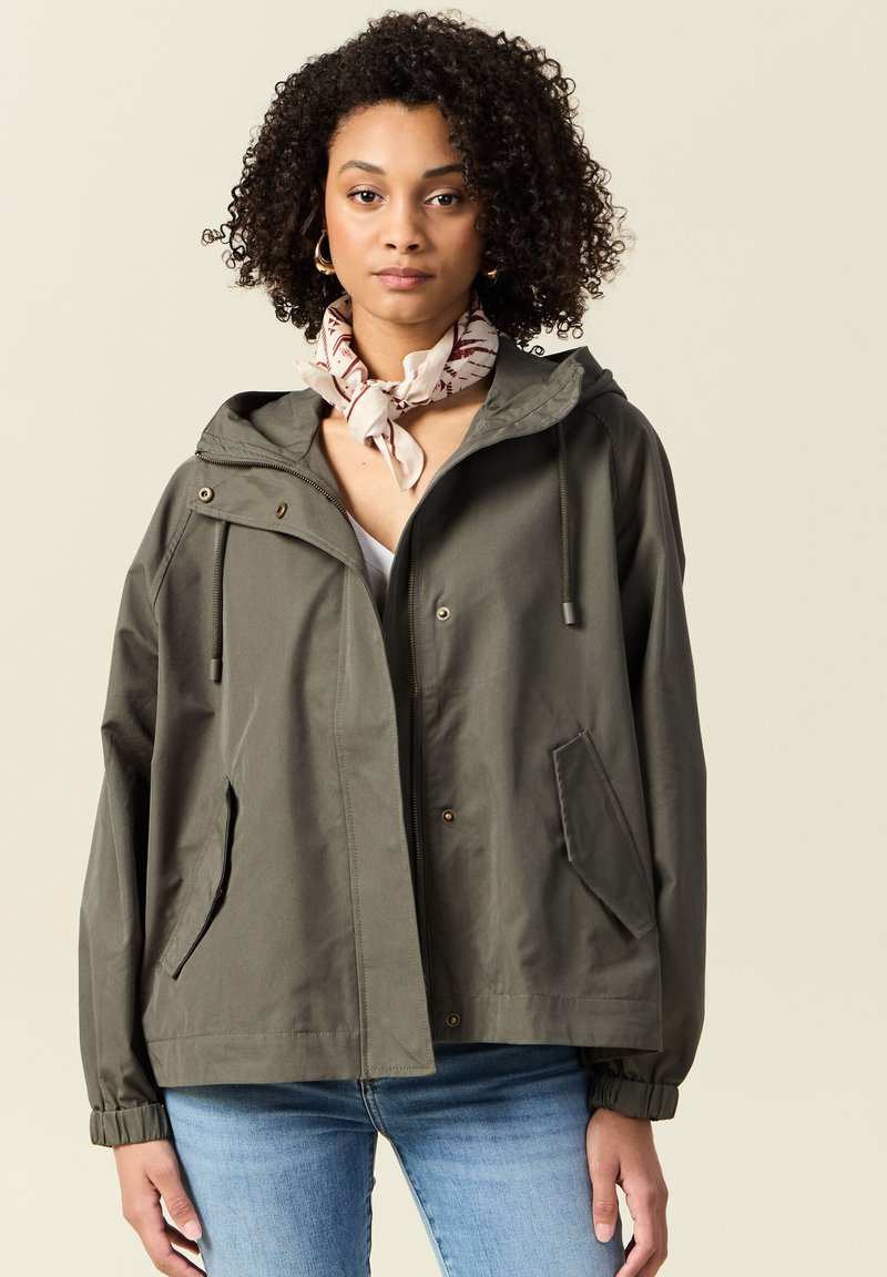 Giacca impermeabile verde oliva con cappuccio, zip frontale, due tasche laterali e polsini elasticizzati. Indossata sopra un top leggero e accessoriate con una sciarpa.