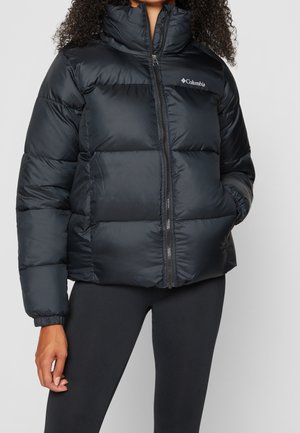 Vrouw draagt een zwarte Columbia donsjas met een hoge kraag en zwarte legging, staand tegen een effen lichtgrijze achtergrond.