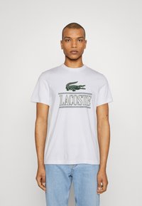 Camiseta de algodón blanca con logo verde de Lacoste y gráfico de cocodrilo en el frente. Con mangas cortas y cuello redondo. Textura ligera y suave.