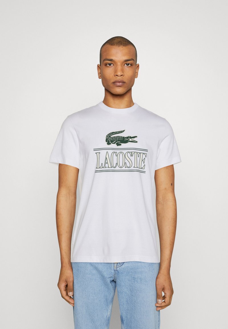 Camiseta de algodón blanca con logo verde de Lacoste y gráfico de cocodrilo en el frente. Con mangas cortas y cuello redondo. Textura ligera y suave.