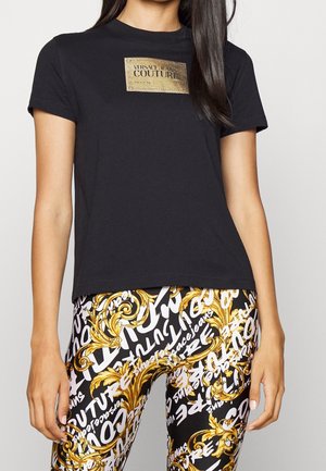 Černé tričko s krátkým rukávem s logem ve zlaté barvě. Kombinováno s legínami s černým, zlatým a bílým vzorem a textem. Hladká struktura látky.
