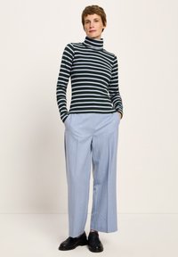 Frau steht mit den Händen in den Taschen, trägt einen schwarz-weiß gestreiften Rollkragenpullover, eine hellblaue weite Hose und schwarze Slipper vor einer weißen Wand.