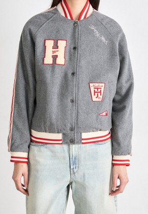 Person iført en grå collegejakke med røde og hvide striber, bogstavet H-mærke og lyseblå jeans, stående mod en hvid baggrund.