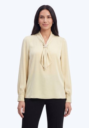 Kvinde med langt mørkt hår iført en lysebeige bluse med lange ærmer, en dekorativ perlebroche og sorte bukser mod en hvid baggrund.