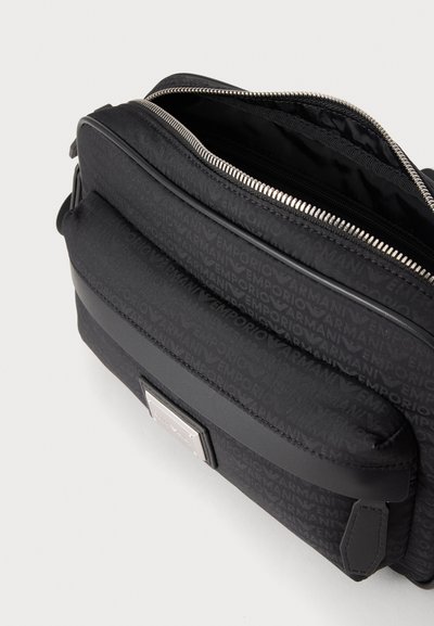 Μαύρη τσάντα Emporio Armani με φερμουάρ, μπροστινή τσέπη και διακοσμητικό σχέδιο στο ύφασμα, σε απλό φόντο.