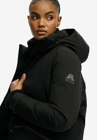 Czarna kurtka puchowa z kapturem, z zapięciem na zamek, miękkim polarem wewnątrz oraz naszywką z logo na lewym rękawie. Materiał jest gładki i wodoodporny.