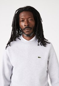 Sweat-shirt gris en coton avec un col rond, arborant un petit logo Lacoste vert sur la poitrine. Le tissu semble doux avec une texture lisse.