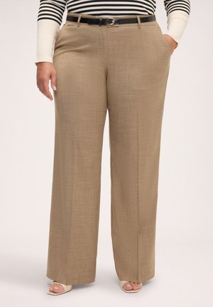 Kvinde iført beige bukser med vidde ben og sort bælte, langærmet stribet top og beige sandaler med åben tå. Hænderne i lommerne.