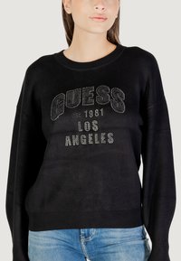 Schwarzer Strickpullover mit rundem Halsausschnitt, ausgestattet mit dem weißen, genoppten "GUESS"-Logo und dem Text "LOS ANGELES". Lockere Passform mit geripptem Saum.