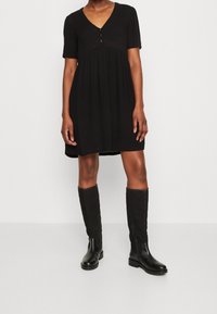 Vestido corto negro de manga corta con escote en V y dos botones en el pecho, silueta fluida, combinado con botas de cuero negras hasta la rodilla.
