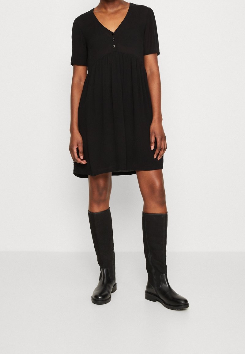 Vestido corto negro de manga corta con escote en V y dos botones en el pecho, silueta fluida, combinado con botas de cuero negras hasta la rodilla.