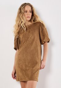 Robe marron en suédine avec des manches courtes bouffantes et un col rond. Présente des coutures verticales pour une silhouette ajustée et des fentes sur les côtés.