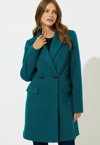 Cappotto verde acqua doppiopetto con risvolti, due tasche frontali e bottoni neri. Texture liscia e design aderente, che termina a metà coscia.