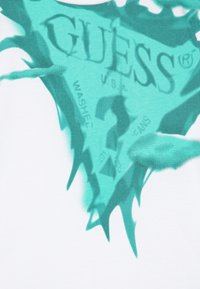 Valkoinen kangas, jossa on suuri turkoosi grafiikka, jossa lukee "GUESS", ja pistekuvio, jossa on roiskefektin ja hienovaraisen tekstin.