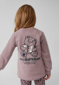 Jasnofioletowy sweatshirt z grafiką misia na plecach, zdobiony srebrnymi akcentami i tekstem. Długie rękawy oraz okrągły dekolt.