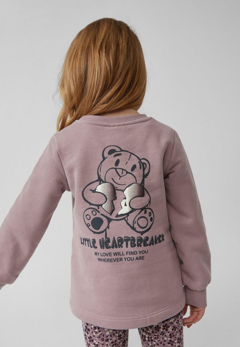 Jasnofioletowy sweatshirt z grafiką misia na plecach, zdobiony srebrnymi akcentami i tekstem. Długie rękawy oraz okrągły dekolt.